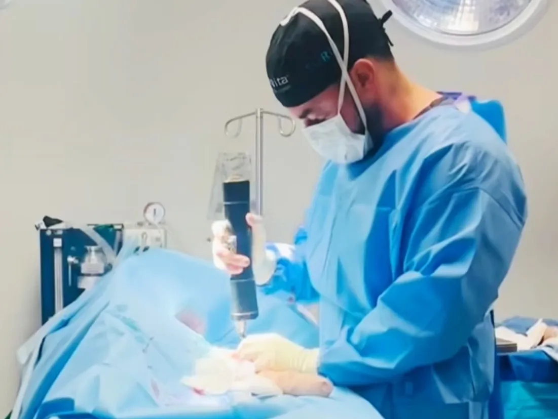 A surgeon in scrubs and a mask using a specialized surgical tool during a procedure in an operating room.