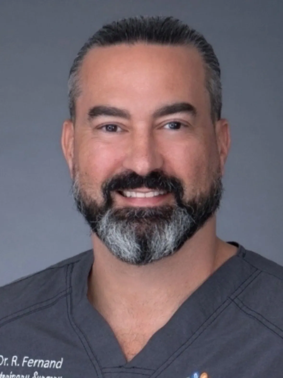 Headshot of smiling Dr R Fernand with a beard wearing gray scrubs against a solid gray background.