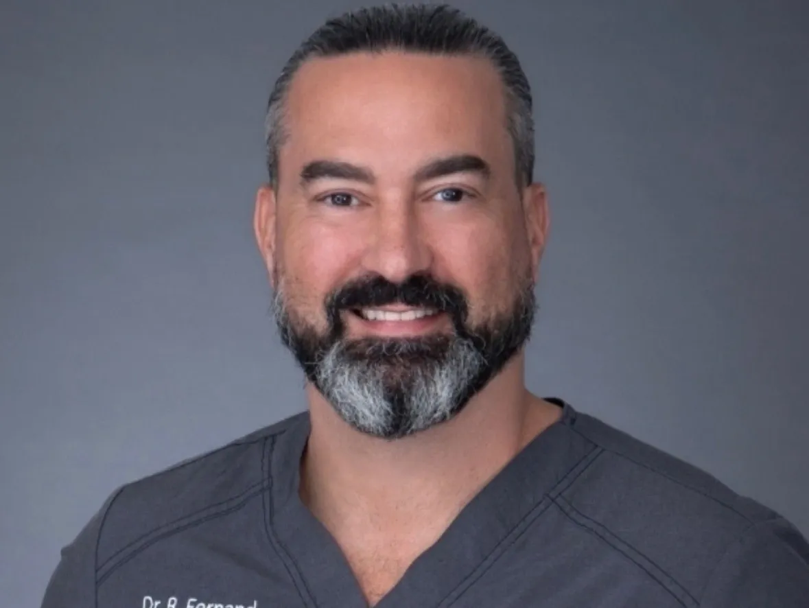 Headshot of a smiling Dr. R. Fernand with a beard, wearing gray scrubs against a solid gray background.