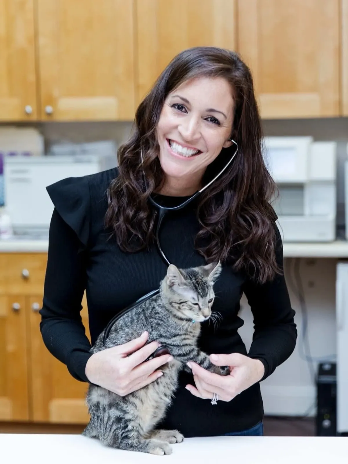 A smiling female veterinarian holds a small tabby kitten while using a stethoscope to examine its heartbeat.