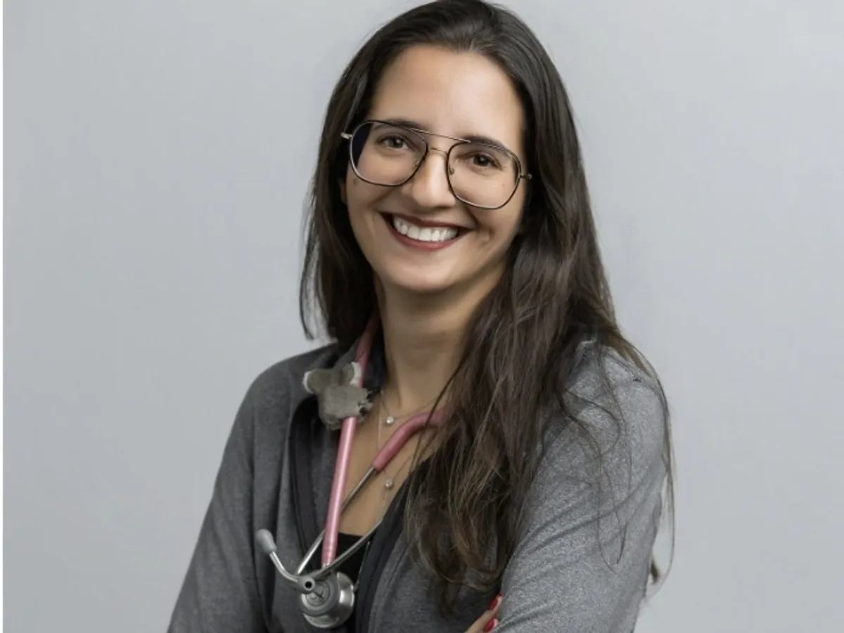 Smiling woman with long brown hair and glasses, wearing a gray top and a pink stethoscope around her neck.