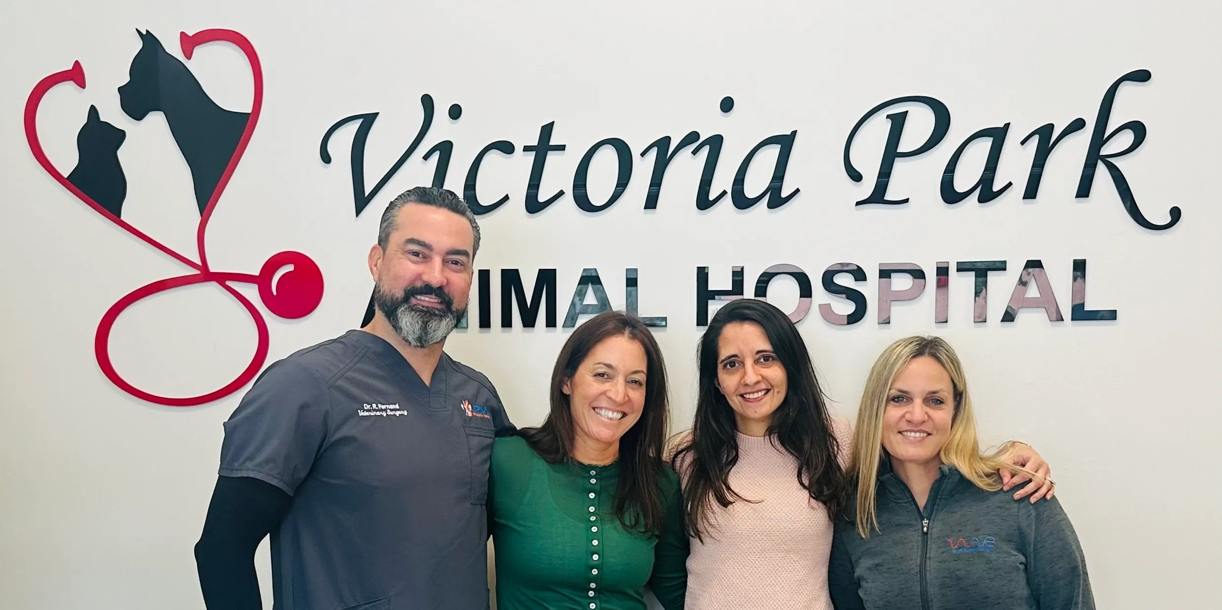 Four smiling staff members posing together in front of a Victoria Park Animal Hospital wall sign.