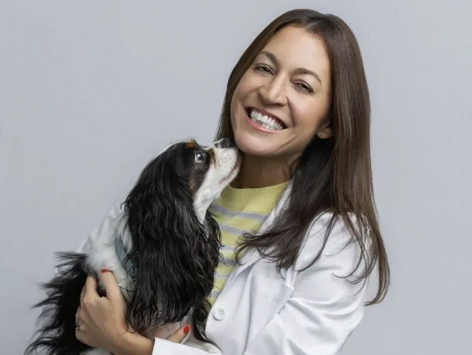 A smiling woman in a white lab coat holding a Cavalier King Charles Spaniel that is nuzzling her chin.