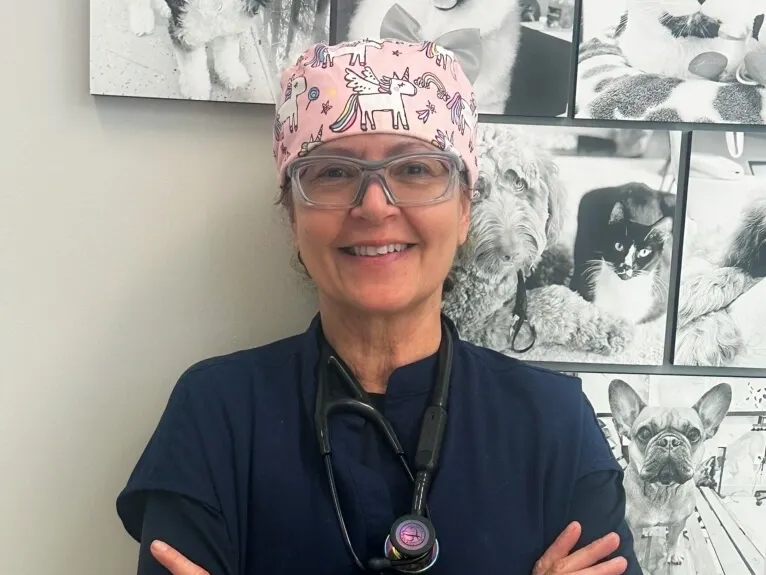 Smiling woman in dark scrubs and a pink unicorn cap, standing in front of a wall of pet photos.