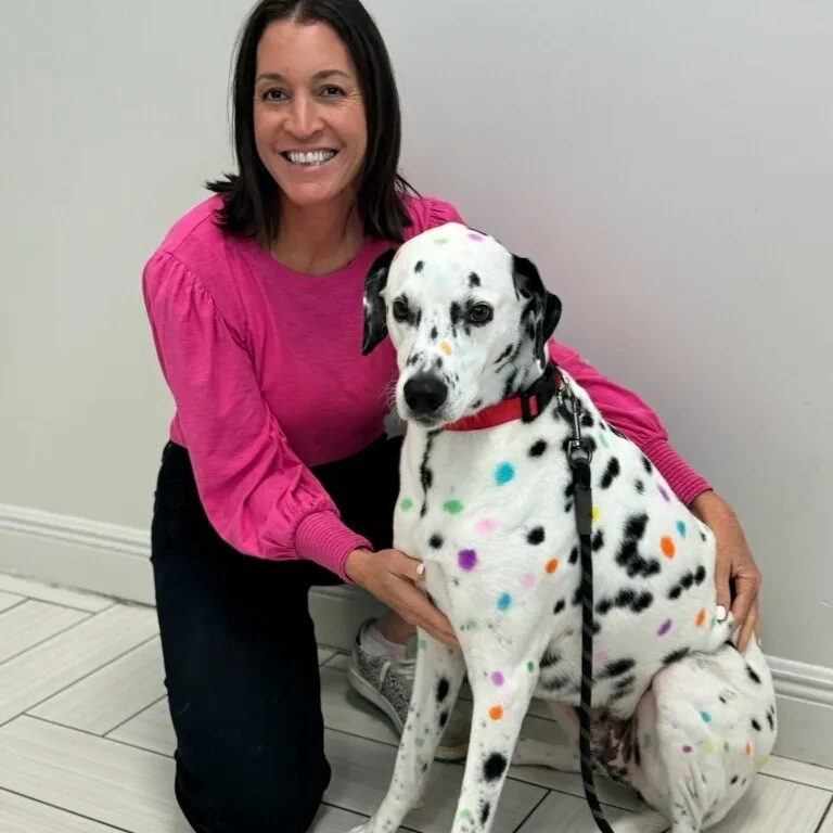 A smiling woman in a pink top kneeling next to a Dalmatian dog covered in colorful, painted spots.