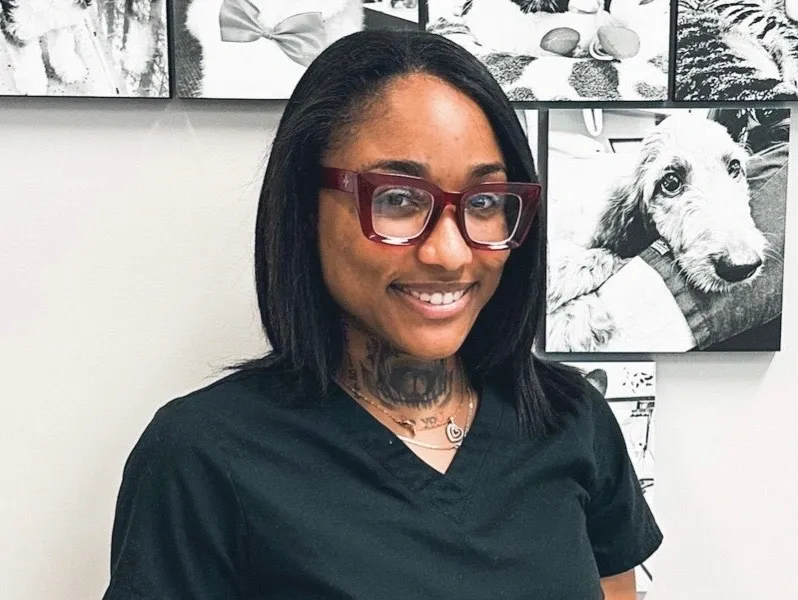 A smiling woman in black scrubs and red glasses, standing in front of a wall with black and white pet photos.