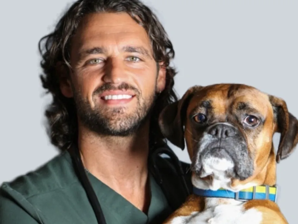 A smiling male veterinarian with curly hair and a beard, wearing green scrubs next to a boxer dog.